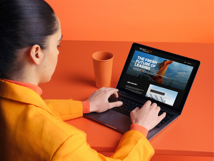 Lady with dark brown hair wearing an orange jacket sitting with her hands on a laptop with the select lease by mobilize website on the screen and an orange background